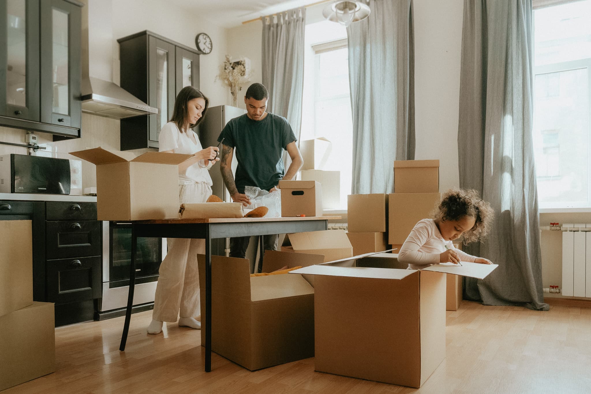 En familj packar inför flytten i sitt kök, medan dottern lekfullt gömmer sig inne i en flyttkartong.