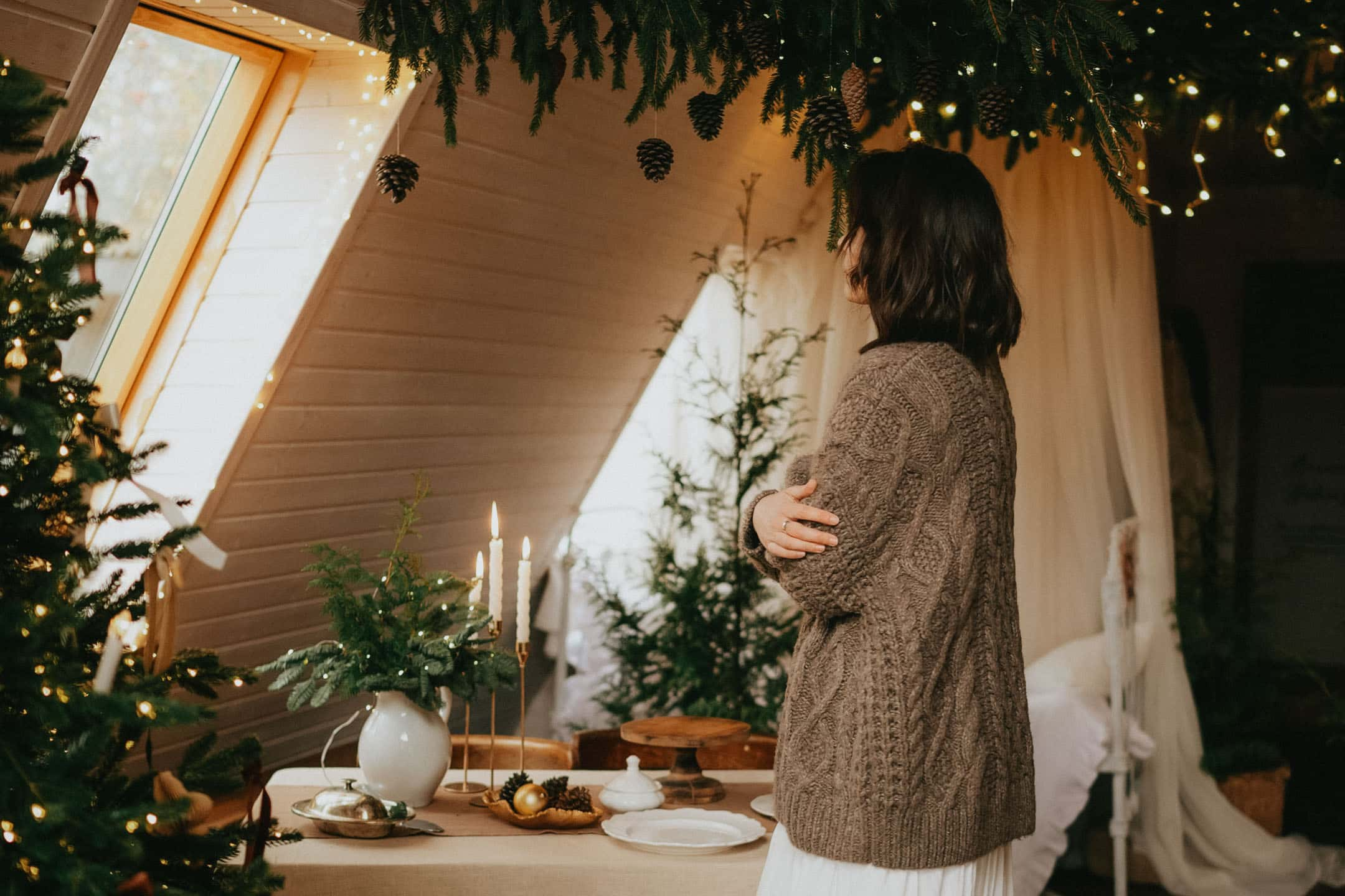 En kvinna i en stickad tröja står i ett mysigt inrett rum med juldekorationer, inklusive en julgran, levande ljus och grankvistar. Ett bord är dukat med rustik julkänsla, och rummet har ett lutande tak med hängande kottar och ljusslingor.