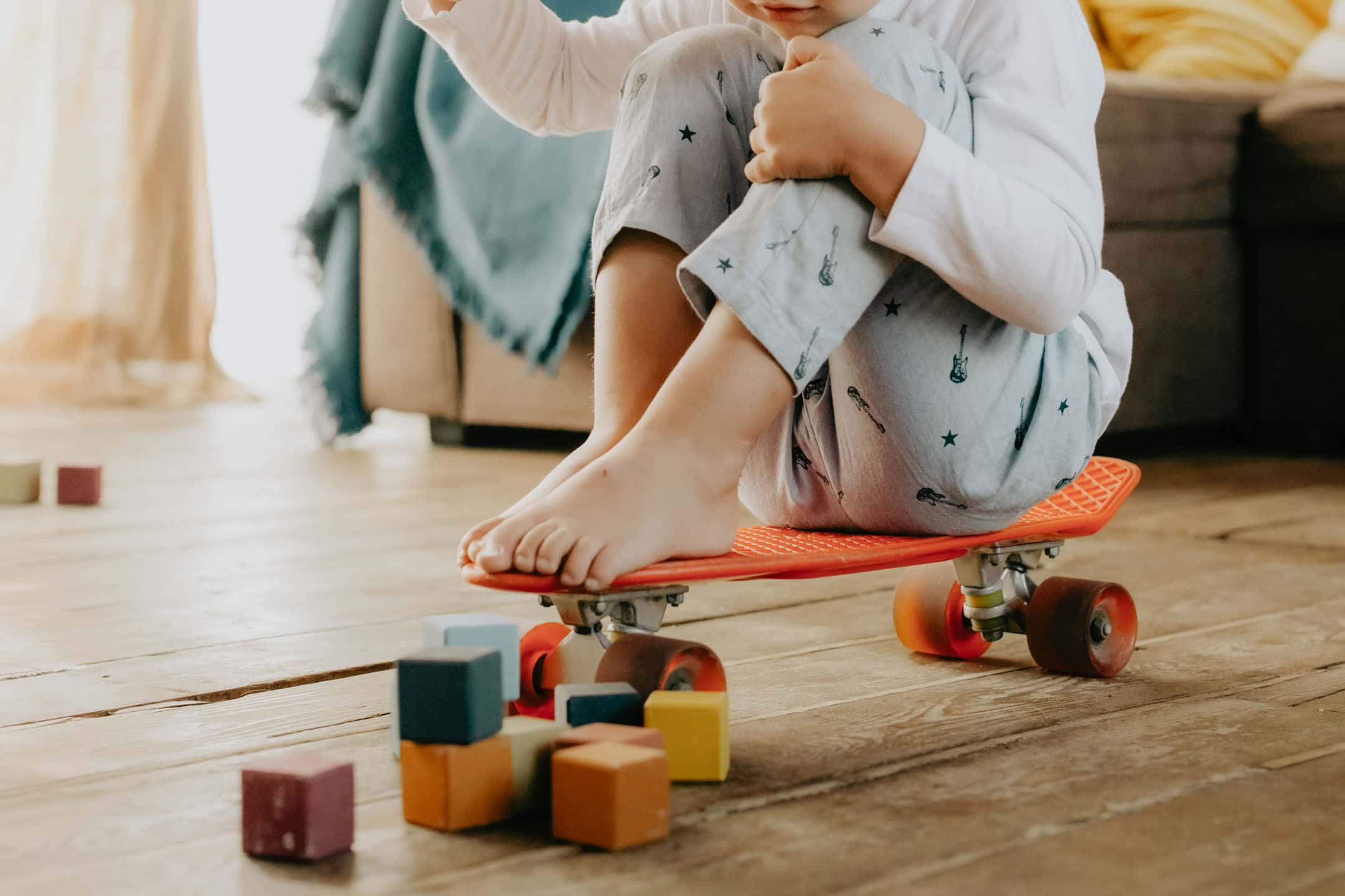 Ett barn i pyjamas sitter på en röd skateboard på ett trägolv, omgiven av färgglada byggklossar. En soffa med en filt syns i bakgrunden.