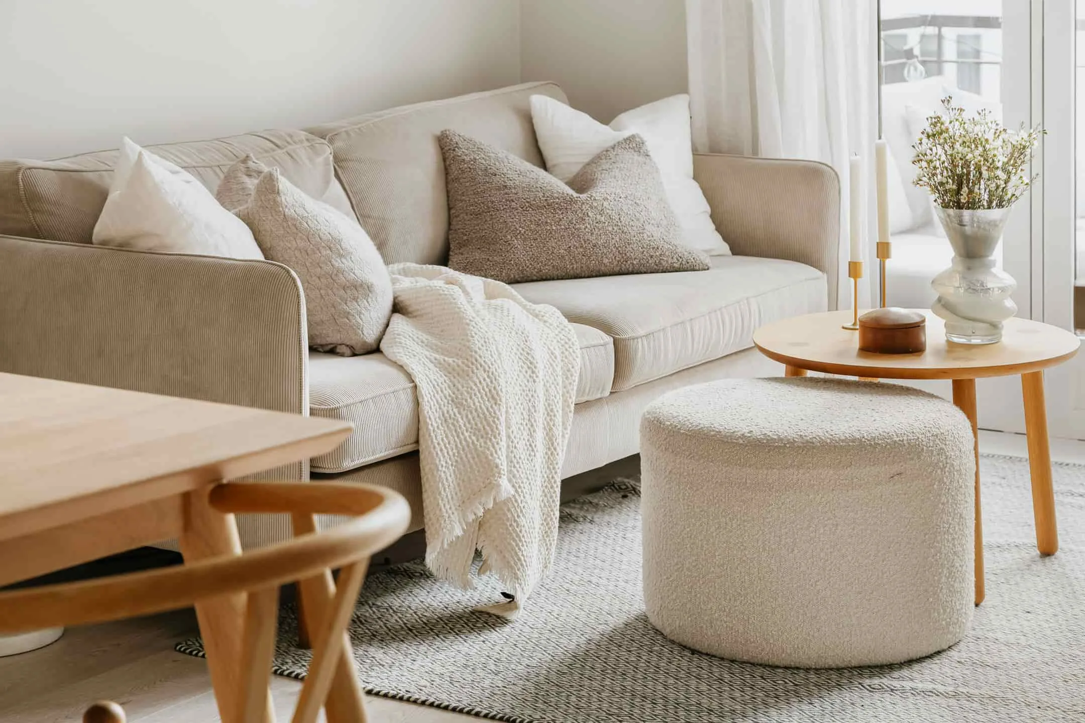 Mysigt och inbjudande vardagsrum med en beige soffa, mjuka kuddar, en stickad pläd och ett runt träbord med ljus och en vas med blommor. Naturligt ljus strömmar in genom fönstret och skapar en harmonisk atmosfär.